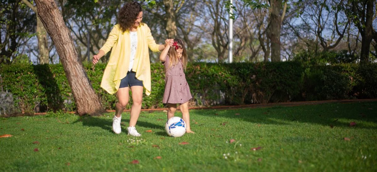 A mother and daughter enjoying a sunny day playing soccer in a lush green park.